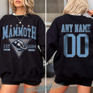 Puede incluir: Sudadera negra con "UTAH MAMMOTH" en letras azul claro y un logotipo del equipo. La parte trasera presenta "ANY NAME" y el n&uacute;mero "00" en azul claro. La sudadera tiene cuello redondo blanco.