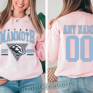 Puede incluir: Sudadera rosa claro con "UTAH MAMMOTH" en letras azul claro. La parte delantera presenta un logotipo del equipo dentro de un tri&aacute;ngulo, con "EST 2024" debajo. La parte trasera muestra "ANY NAME" y el n&uacute;mero "00" en azul claro.