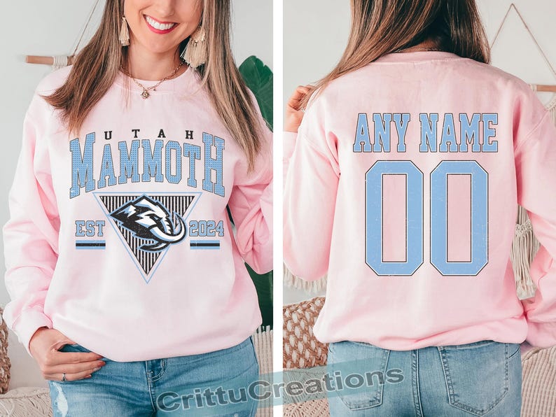 Puede incluir: Sudadera rosa claro con "UTAH MAMMOTH" en letras azul claro. La parte delantera presenta un logotipo del equipo dentro de un tri&aacute;ngulo, con "EST 2024" debajo. La parte trasera muestra "ANY NAME" y el n&uacute;mero "00" en azul claro.