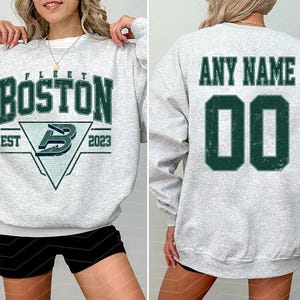 May include: A light gray sweatshirt with the word "BOSTON" in dark green block letters. Below the word "BOSTON" is a triangle with the letter "B" inside. The back of the sweatshirt has the words "ANY NAME" above the number "00".