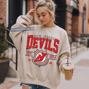 Może przedstawiać: Beżowa bluza z napisem "NEW JERSEY DEVILS" w kolorze czerwonym, ze efektem postarzenia. Bluza prezentuje również logo drużyny i rok "1974". Rękawy są długie, a ogólny styl jest swobodny.