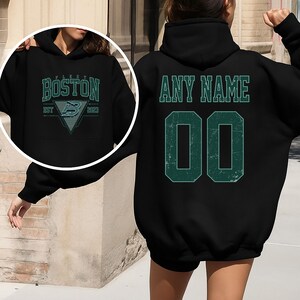 May include: Black hoodie with teal lettering and graphics. The front features "BOSTON" above a triangle with a stylized "B" and the text "EST 2023". The back displays "ANY NAME" above the number "00" in teal.