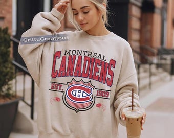 Puede incluir: Sudadera beige con el texto "MONTREAL CANADIENS" en rojo, con el logotipo del equipo. La sudadera tiene un aspecto vintage y desgastado. El año "1909" está impreso debajo del logotipo. La sudadera es de estilo casual.