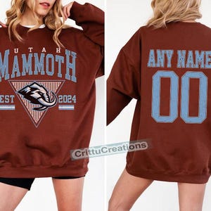 Puede incluir: Sudadera granate con el texto "UTAH MAMMOTH" en azul claro, un logotipo de mamut y "EST 2024". La parte trasera muestra "ANY NAME" y el n&uacute;mero "00" en azul claro. La sudadera tiene cuello redondo y es una prenda informal.