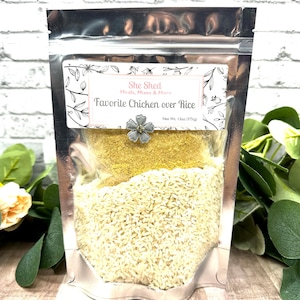 May include: A silver pouch with a white label that reads "She Shed Meals, Mixes & More Favorite Chicken over Rice Net Wt. 13oz/375(g)". The pouch contains white rice and a yellow powder.
