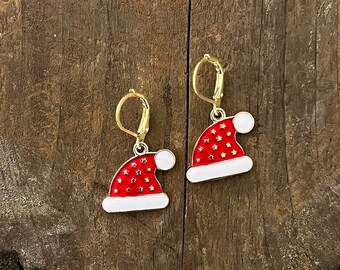 Puede incluir: Un par de pendientes navideños. Cada pendiente presenta un diseño de gorro de Papá Noel rojo con ribete blanco y cierres de palanca dorados. Los gorros están decorados con pequeñas formas de estrella.