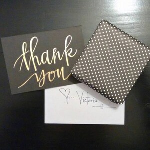 May include: A black card with gold lettering that says "thank you". A white card with a heart and the handwritten text "...Victoria" is next to the black card. A black and white polka dot gift box is in the background.
