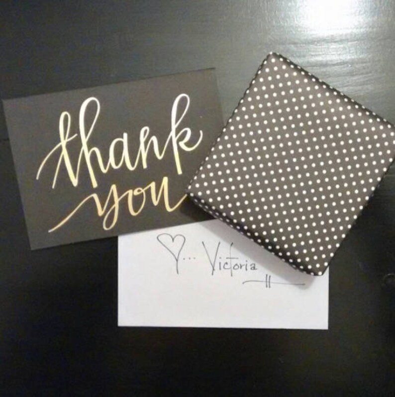 May include: A black card with gold lettering that says "thank you". A white card with a heart and the handwritten text "...Victoria" is next to the black card. A black and white polka dot gift box is in the background.