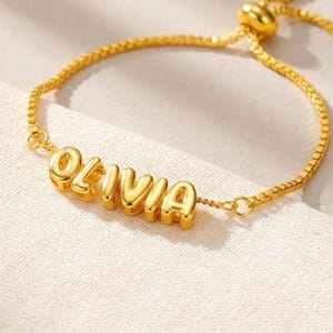May include: A gold-toned adjustable bracelet featuring a box chain and a central nameplate with the letters O-L-I-V-I-A in a rounded, three-dimensional font. The bracelet includes a sliding bead mechanism to adjust the fit of the chain.