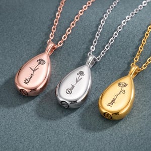 May include: Three teardrop-shaped pendant necklaces in rose gold, silver, and gold tones. Each pendant features a delicate engraved flower and a name: Klaus, Dad, and Papa. The pendants hang from matching metal chains with a screw-in base.