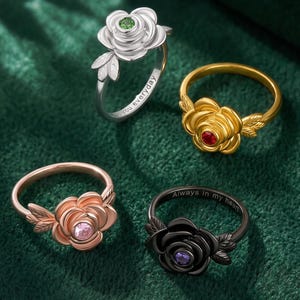 May include: Four metal rings shaped like roses with leaves. One silver ring with a green stone, one gold ring with a red stone, one rose gold ring with a pink stone, and one black ring with a purple stone. The silver ring says you everyday.