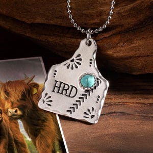 May include: A silver-colored pendant necklace with a turquoise stone and the letters "HRD". The pendant has a textured surface with decorative patterns. A photo of two brown cows is in the background.