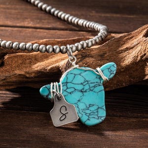 May include: A turquoise cow pendant necklace with a silver-tone chain. The pendant is wire-wrapped and features a turquoise stone with black veining. A small, silver-tone tag with the letter "S" hangs below the pendant. The necklace is displayed on a wooden surface.