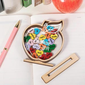 Könnte beinhalten: Holz-Andenkenbox in Apfelform mit klarer Vorderseite, gefüllt mit bunten herzförmigen Namensschildern. Die Box trägt den Text "NGUINE 2020-2024". Ein rosa Stift und ein Holzständer sind ebenfalls sichtbar. Thema: Geschenk zur Lehreranerkennung.