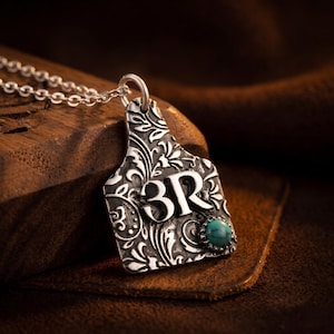 May include: A silver-toned pendant shaped like a livestock ear tag, featuring an intricate embossed floral pattern and the letters 3R in relief. A small, round, blue-green turquoise stone is set in the lower right corner, attached to a silver chain.