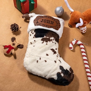 May include: A festive Christmas stocking with a cowhide design, a white trim, and the name "JOHN" on a brown leather label. The image includes Christmas decorations such as a gift, baubles, and a candy cane.