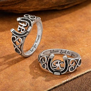 Op de afbeelding: Twee zilverkleurige ringen met hoefijzerontwerpen. Eén ring heeft de tekst "Love You Forever" en de andere luidt "Infinite Luck for You". De ringen hebben zwarte accenten en ingewikkeld krulwerk.