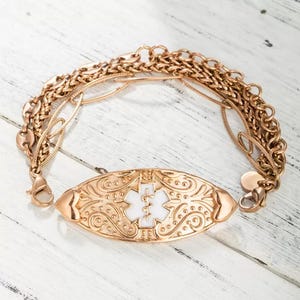 May include: A gold-coloured medical alert bracelet with a white enamel centre displaying the Star of Life symbol. The oval plate is decorated with detailed floral and scrollwork patterns. The bracelet has multiple chain strands.