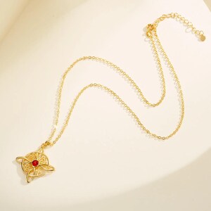 May include: Gold-colored necklace featuring a pendant with a Celtic knot design and a small red gemstone at the center. The chain is delicate and adjustable. The necklace is displayed on a light-colored surface.