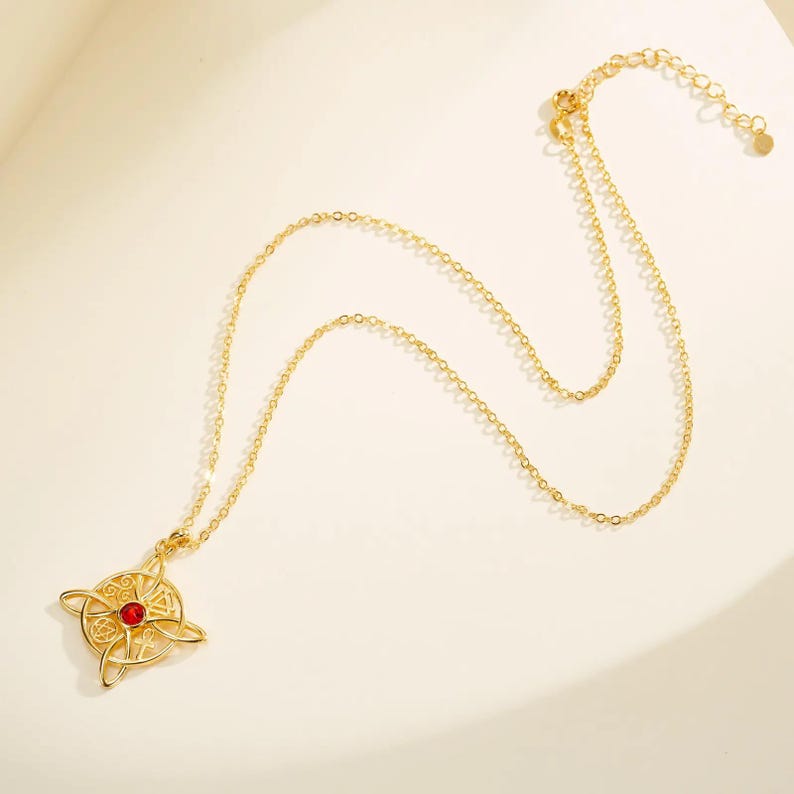 May include: Gold-colored necklace featuring a pendant with a Celtic knot design and a small red gemstone at the center. The chain is delicate and adjustable. The necklace is displayed on a light-colored surface.