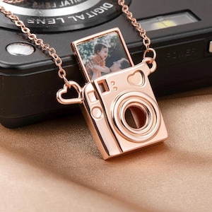 May include: Rose gold camera pendant necklace with a small photo inside. The camera pendant has heart-shaped details and a chain. The necklace is displayed on a black camera.