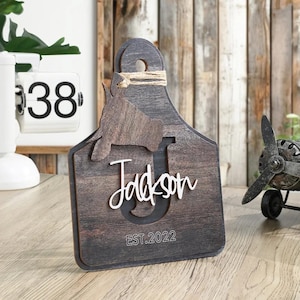 May include: A dark brown wooden tag with the name "Jackson" in white script, a cow silhouette, and the text "EST.2022". The tag has a jute twine accent and a lighter brown cow silhouette.