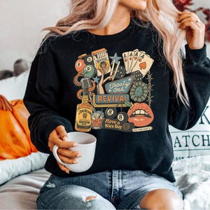 Peut inclure: Sweat-shirt noir &agrave; col rond avec un motif r&eacute;tro. Le motif comprend une bouteille de bourbon Jim Beam, des cartes &agrave; jouer, un microphone et les mots "Treaty Oak Revival" et "Have a Nice Day".