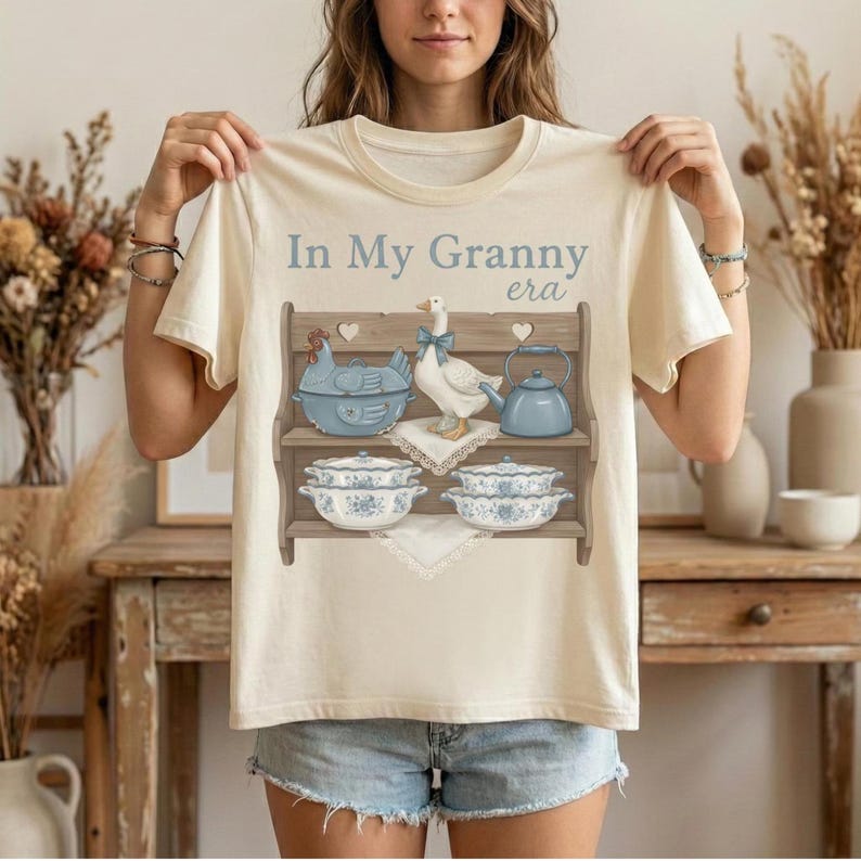 May include: Cream-colored t-shirt with the text "In My Granny era" above a graphic of a shelf with a blue chicken, goose, teapot, and floral bowls. The shirt is being held up by a person.