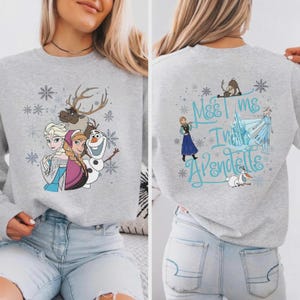 Peut inclure: Un sweat-shirt gris avec des personnages du film d'animation "La Reine des Neiges". Le devant montre Elsa, Anna, Olaf et Sven, avec des flocons de neige. Le dos porte l'inscription "Meet me in Arendelle" avec d'autres personnages et flocons.