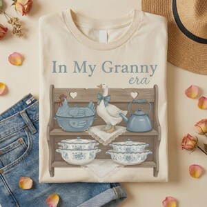 May include: Cream-colored t-shirt featuring a graphic of a wooden shelf with a blue chicken-shaped casserole dish, a white goose with a blue bow, and blue floral bowls. The text "In My Granny era" is printed above the shelf.