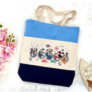 May include: A blue, white, and navy blue canvas tote bag with a design of Mickey Mouse, Donald Duck, Goofy, Pluto, and Minnie Mouse in porthole frames. The design includes nautical elements like anchors, seashells, and an octopus.