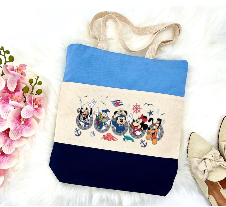 May include: A blue, white, and navy blue canvas tote bag with a design of Mickey Mouse, Donald Duck, Goofy, Pluto, and Minnie Mouse in porthole frames. The design includes nautical elements like anchors, seashells, and an octopus.