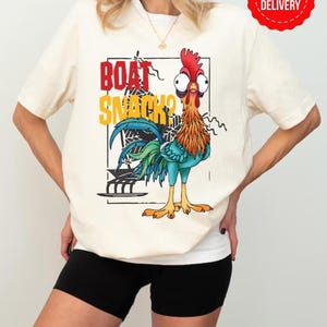 Puede incluir: Camiseta color crema con un gallo de dibujos animados y el texto "BOAT SNACKS?" en rojo y amarillo. El gallo tiene una cresta roja, plumas naranjas y azules y ojos grandes. Una pegatina roja de "FAST DELIVERY" es visible.