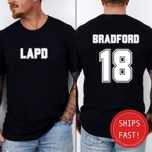 May include: Black t-shirt with white text. The front of the shirt reads "LAPD" in a bold, block font. The back of the shirt displays "BRADFORD" above the number "18" in a similar font. The shirt is a crew neck.