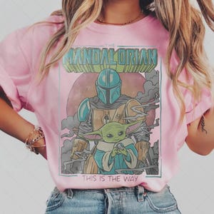 Puede incluir: Camiseta rosa claro con un dise&ntilde;o de The Mandalorian y Grogu, con el texto "THE MANDALORIAN" y "THIS IS THE WAY". El dise&ntilde;o incluye una figura con casco y un peque&ntilde;o personaje verde, sobre un fondo rosa.