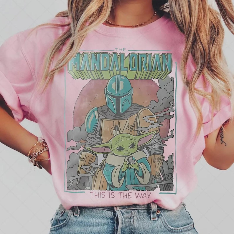 Puede incluir: Camiseta rosa claro con un dise&ntilde;o de The Mandalorian y Grogu, con el texto "THE MANDALORIAN" y "THIS IS THE WAY". El dise&ntilde;o incluye una figura con casco y un peque&ntilde;o personaje verde, sobre un fondo rosa.