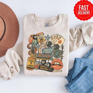 Peut inclure: Sweat-shirt cr&egrave;me avec un motif color&eacute; comprenant une bouteille de bourbon, des cartes &agrave; jouer, un microphone et le texte "Treaty Oak Revival". Le motif comprend &eacute;galement le texte "Have a Nice Day" et "No Vacancy".