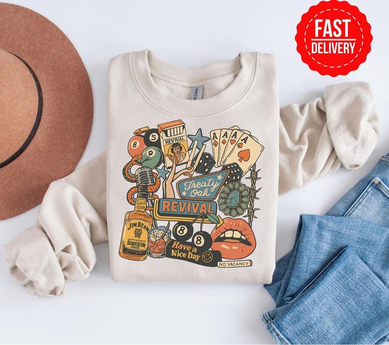 Peut inclure: Sweat-shirt cr&egrave;me avec un motif color&eacute; comprenant une bouteille de bourbon, des cartes &agrave; jouer, un microphone et le texte "Treaty Oak Revival". Le motif comprend &eacute;galement le texte "Have a Nice Day" et "No Vacancy".