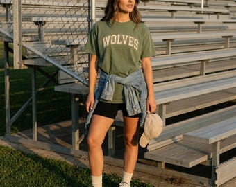 May include: An olive green t-shirt with the word WOLVES printed in white block letters across the chest, paired with black biker shorts, a light blue denim jacket tied around the waist, white sneakers with green accents, and a beige baseball cap.