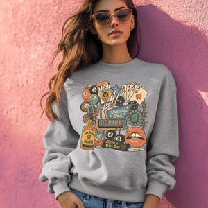 Peut inclure: Un sweat-shirt gris orn&eacute; d'un motif color&eacute; d'inspiration vintage. Le design comprend une bouteille de Jim Beam, des cartes &agrave; jouer, des d&eacute;s, un microphone et les mots "Treaty Oak Revival" et "Have a Nice Day".