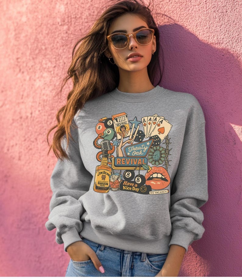 Peut inclure: Un sweat-shirt gris orn&eacute; d'un motif color&eacute; d'inspiration vintage. Le design comprend une bouteille de Jim Beam, des cartes &agrave; jouer, des d&eacute;s, un microphone et les mots "Treaty Oak Revival" et "Have a Nice Day".