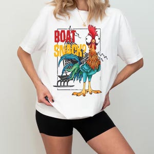 Puede incluir: Camiseta blanca con un dise&ntilde;o de gallo de dibujos animados y el texto "BOAT SNACK?" en rojo y amarillo. El gallo tiene una cresta roja, plumas naranjas y azules y ojos grandes. Camiseta casual y unisex.