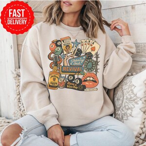 Peut inclure: Sweat-shirt beige avec un motif color&eacute; repr&eacute;sentant une bouteille de bourbon Jim Beam, des cartes &agrave; jouer, des d&eacute;s et les mots "Treaty Oak Revival" et "Have a Nice Day". Le motif comprend des l&egrave;vres et un panneau "No Vacancy".