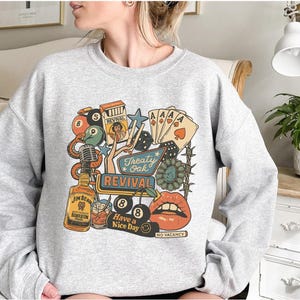 Peut inclure: Un sweat-shirt gris clair avec un motif graphique color&eacute;. Le motif comprend une bouteille de bourbon Jim Beam, des cartes &agrave; jouer, des d&eacute;s, un microphone et les mots "Treaty Oak Revival" et "Have a Nice Day".