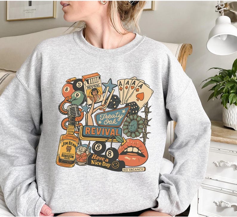 Peut inclure: Un sweat-shirt gris clair avec un motif graphique color&eacute;. Le motif comprend une bouteille de bourbon Jim Beam, des cartes &agrave; jouer, des d&eacute;s, un microphone et les mots "Treaty Oak Revival" et "Have a Nice Day".