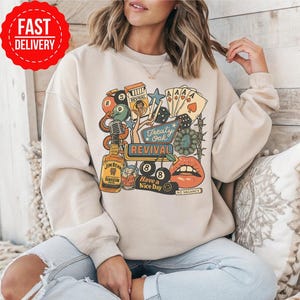 Peut inclure: Sweat-shirt beige avec un motif color&eacute;. Le motif comprend une bouteille de bourbon Jim Beam, des cartes &agrave; jouer, des d&eacute;s et les mots "Treaty Oak Revival" et "Have a Nice Day". Le motif comprend une paire de l&egrave;vres et une boule de billard.