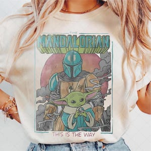 Puede incluir: Camiseta color crema con un gr&aacute;fico de The Mandalorian y Grogu. El dise&ntilde;o incluye el texto "THE MANDALORIAN" en una fuente retro, con "THIS IS THE WAY" debajo. La obra de arte muestra a los personajes en una ilustraci&oacute;n estilizada tipo caricatura.