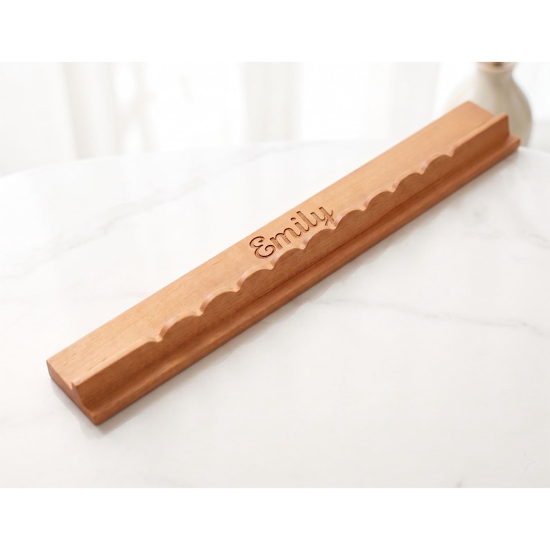 May include: A wooden Scrabble tile holder with a scalloped edge design. The holder is light brown and features the name "Emily" engraved in a cursive font. The holder is designed to hold Scrabble tiles and is made of wood.