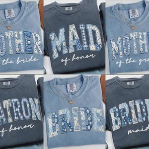 May include: A collection of blue crewneck sweatshirts with various wedding-related titles. The sweatshirts feature patchwork lettering with floral and gingham patterns. Titles include "Mother of the Bride," "Maid of Honor," and "Bridesmaid."