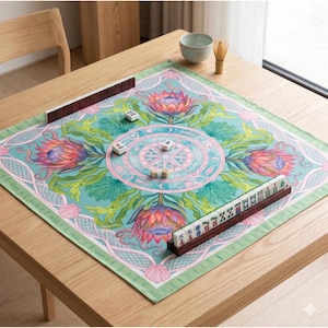 May include: A square Mahjong game mat with a floral design in shades of teal, pink, and green. The mat features a central compass rose design and is surrounded by decorative borders. Brown tile racks and game tiles are arranged on the mat.